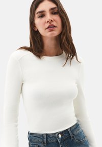 Donna con lunghi capelli castani, che indossa una maglietta attillata bianca a maniche lunghe e jeans blu a vita alta, in piedi contro uno sfondo neutro.
