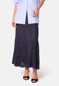 Marineblauer, gefalteter Maxirock mit metallischen Fäden, weicher Textur und ausgestelltem Saum. Kombiniert mit einem hellblauen Hemd und schwarzen Offen-Zehen-Absätzen.