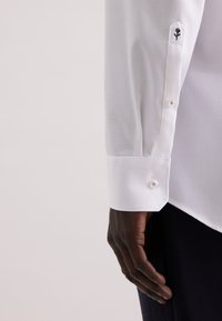 Mano di carnagione scura accanto a un polsino di camicia a maniche lunghe di colore bianco con bottoni e una piccola ricamo di rosa nera vicino al polso.