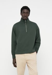 Mørkegrøn zip-up sweatshirt med høj krave, afslappet pasform og ribbede manchetter, parret med beige bukser. Blødt stof, minimalistisk design.
