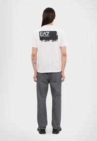 Balts īsām piedurknēm T-krekls ar melnu kvadrātu grafiku aizmugurē, kurā ir teksts "EA7 Emporio Armani". Kombinēts ar pelēkiem platām biksēm.