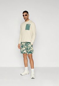 Langärmeliges cremefarbenes Hemd mit grünem Brusttasche, kombiniert mit gemusterten grünen und beigefarbenen Shorts und weißen Turnschuhen. Runde Sonnenbrille.