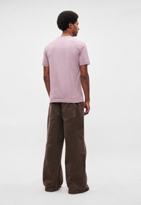 Άντρας με χτένισμα αφρο φορώντας ένα λιλά κοντομάνικο πουκάμισο και φαρδιά καφέ παντελόνια στέκεται με την πλάτη προς τον θεατή πάνω σε απλό φόντο.