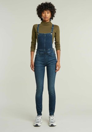 Denim overall met een fitted pasvorm, donkerblauwe kleur, verstelbare banden en voorkruisen; gecombineerd met een olijfgroene coltrui met lange mouwen.