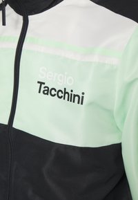 Svart och mintgrön träningsjacka med nätstruktur. Har vita horisontella ränder och svart text "Sergio Tacchini" på framsidan.