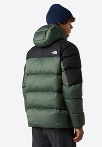 Grön och svart puffjacka med huva. Har en glänsande yta, tjock stoppning och en logga på övre delen av ryggen. Ribbade blå mössa synlig.