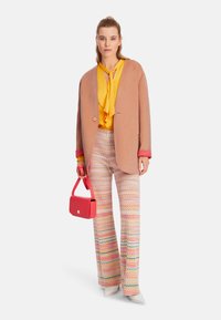 Hellrosa Wollblazer, leuchtend gelbe Bluse, gemusterte weite Hose und eine kleine rote Handtasche. Solide Schuhe vervollständigen das Ensemble.