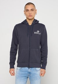 Marineblauwe zip-up hoodie gemaakt van zachte stof, voorzien van een capuchon met trekkoord, voorzakken en een wit "REPLAY" logo op de linkerbovenkant van de borst.