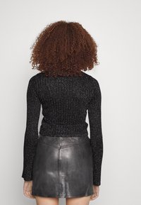 Kvinna med lockigt brunt hår som bär en glittrig svart långärmad topp och en svart läderminikjol, stående vänd mot en vit vägg.