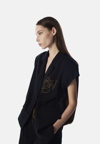 Czarna bezrękawnik z złotym haftem "S W." Posiada wyłogi, przednie kieszenie oraz luźny krój.