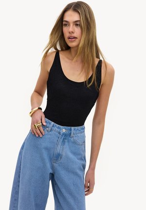 Femme aux longs cheveux portant un haut noir sans manches en dentelle et un jean bleu ample taille haute, accessoirisée avec des bagues et un bracelet en or.