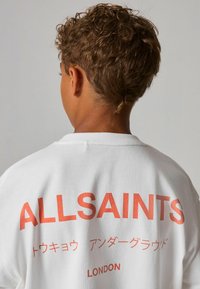 Bílá oversized tričko s velkým červeným textem "ALLSAINTS" na zádech, s japonskými znaky a "LONDON" pod tím. Měkká látka.