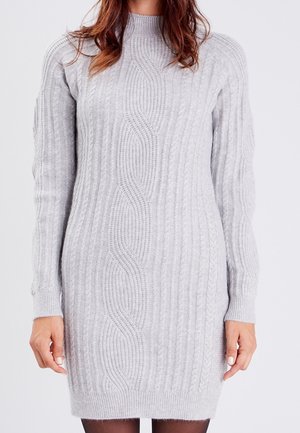 Femme portant une robe-pull en maille torsadée gris clair à manches longues, col montant, et des collants noirs.