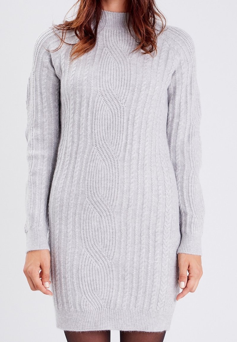 Femme portant une robe-pull en maille torsadée gris clair à manches longues, col montant, et des collants noirs.