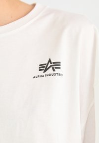 Valkoinen puuvillatee paita, jossa on musta logo tyylitellyllä "A":lla ja "Alpha Industries" -tekstillä vasemmassa rinta-alueessa. Pyöreä kaula-aukko.