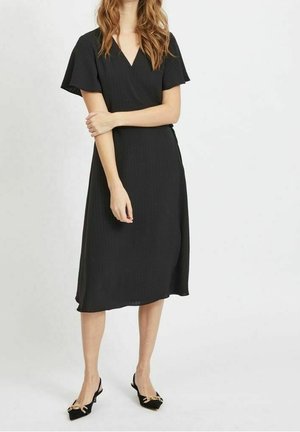 Kobieta stojąca w czarnej, owijanej sukience sięgającej kolan z falbanowymi rękawami oraz czarnych, szpiczastych butach na obcasie z odkrytą piętą i złotym łańcuszkiem jako ozdobą.