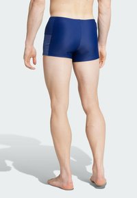 Bañador azul con una textura suave y brillante; presenta una parte trasera sólida y paneles laterales con rayas claras; longitud hasta la mitad del muslo.