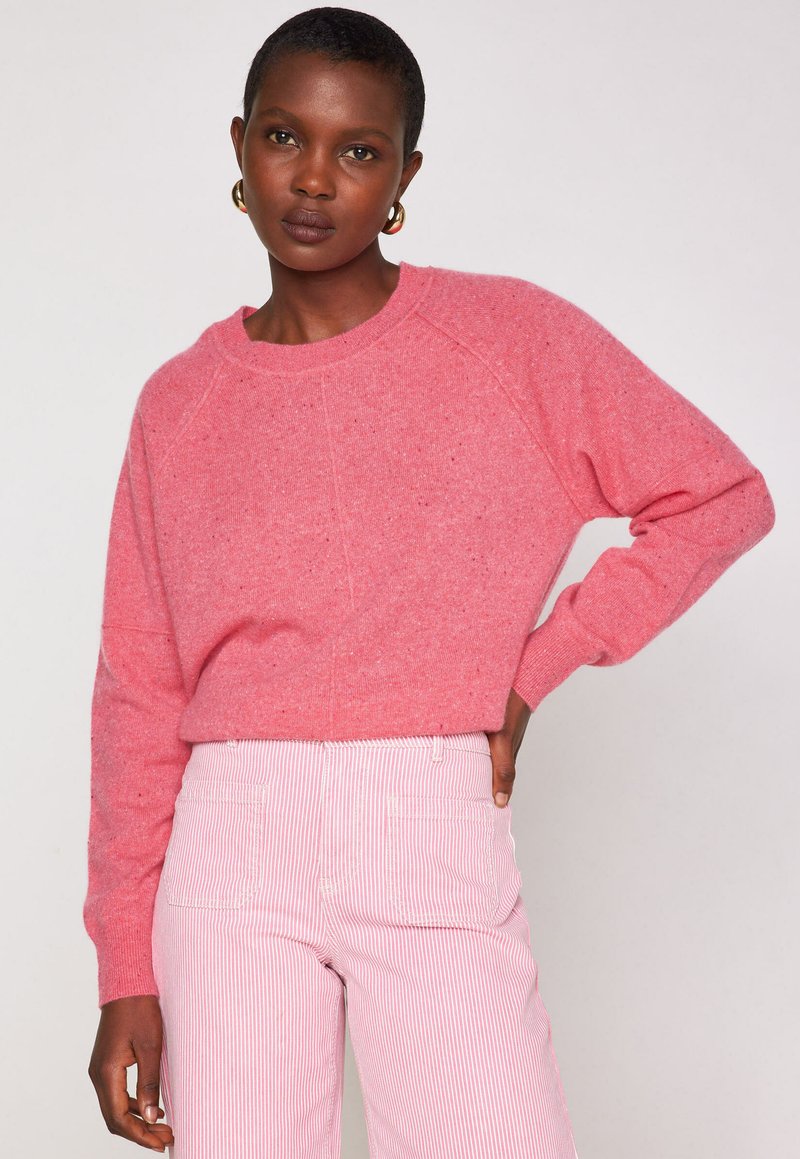 Donna con capelli corti, che indossa orecchini a cerchio dorati, un maglione rosa a girocollo e pantaloni a righe rosa e bianchi, in piedi con una mano sulla vita.