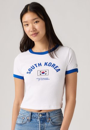 Mladá žena s dlhými tmavými vlasmi, oblečená v bielom krátkom tričku s modrými okrajmi, textom „South Korea“ a vlajkou, v kombinácii s modrými džínmi.