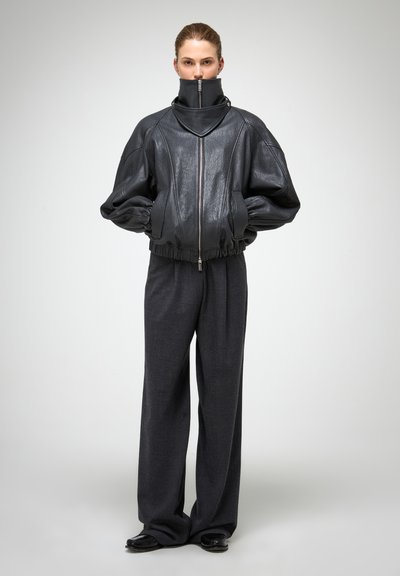 Schwarze Lederbomberjacke mit hohem Kragen, Reißverschluss vorne und gerafften Bündchen, kombiniert mit locker sitzenden, dunkelgrauen Hosen.