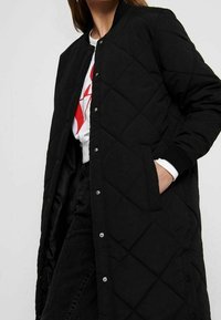 Czarny pikowany długi płaszcz z zapięciem na zatrzaski, bocznymi kieszeniami i ściągaczami, o gładkiej fakturze i stonowanym designie.