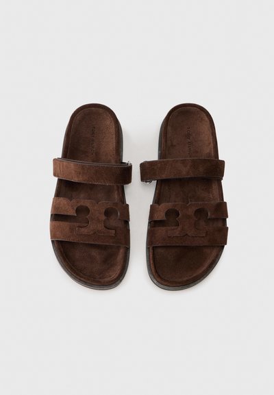 Brown suede slip-on sandals with wide straps, featuring cut-out patterns and a flat sole on a white background.