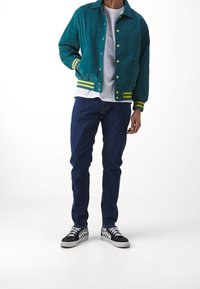 Teal corduroy jacket with yellow and green striped cuffs, paired with a white shirt and blue jeans. Black and white checkered sneakers.