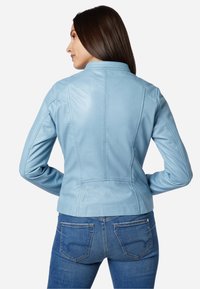 Himmelblaues Lederjacke mit tailliertem Design, Stehkragen und Nahtdetails. Kombiniert mit dunkelblauen Jeans. Glatte Textur.