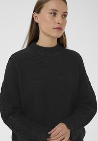 Pull noir côtelé avec un col montant et un tissu texturé. Il possède des détails lacés sur les manches et une coupe décontractée.