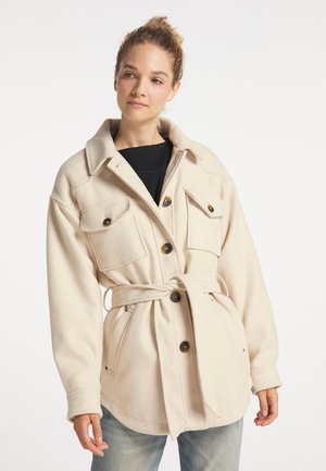 Femme avec les cheveux attachés en chignon portant une veste beige ceinturée avec de gros boutons, des poches poitrine, et un jean bleu clair sur fond blanc.