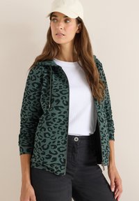 Felpa con zip a motivo leopardato verde, dotata di cappuccio, dettagli neri e maniche lunghe, indossata sopra una T-shirt bianca, abbinata a pantaloni neri.