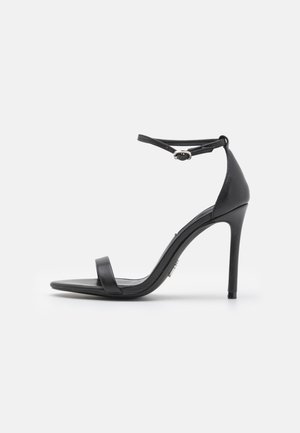 Sandalias de tacón alto negras con punta abierta, tiras delgadas y hebilla en el tobillo. Textura de cuero suave con un diseño de punta afilada.