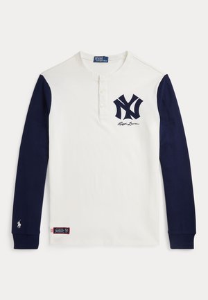 Bílá košile s dlouhým rukávem henley s námořnickými rukávy, logo New York Yankees a podpis Ralph Lauren na prsou, a malý erb koně na manžetě rukávu.