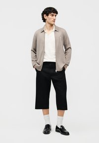 Mladý muž oblečený v béžovom kardigane na zips, bielej košeli, čiernych širokých nohaviciach skracovaných nad členky, bielych ponožkách a čiernych mokasínach, stojaci s rukami v kapsách.