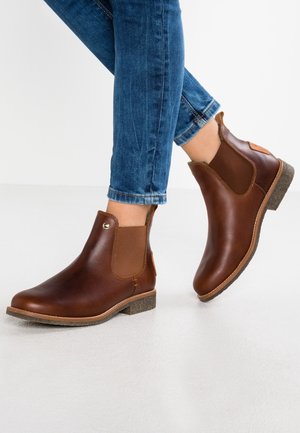 Bottes en cuir marron avec panneaux latéraux élastiques portées avec un jean en denim bleu, montrant le pied droit légèrement levé du sol.