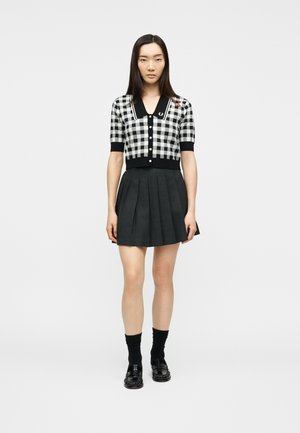 Jeune femme portant un cardigan à manches courtes à carreaux noirs et blancs, une jupe plissée noire, des chaussettes noires et des mocassins noirs, debout devant un fond blanc.