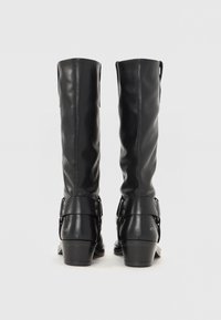 Botas de cuero negro hasta la rodilla con un diseño estructurado, parte superior curva y detalles de hebilla en las correas de los tobillos, con un tacón bajo apilado.