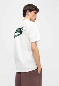 Νέος άνδρας με λευκό μπλουζάκι Nike με πράσινο λογότυπο στο πίσω μέρος, φορώντας καφέ παντελόνια, στέκεται σε προφίλ με το ένα χέρι στην τσέπη.