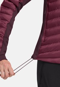 Mano che regola il cordino all'orlo di una giacca trapuntata ma rossa indossata sopra pantaloni neri su uno sfondo semplice.