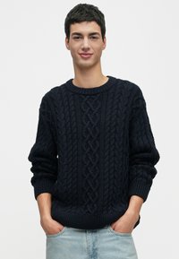 Mlad moški stoji z rokami v žepih, oblečen v temno modro pletivo z nitmi in svetlo modre kavbojke, proti navadni beli ozadju.