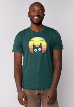 Groene katoenen t-shirt met een centrale afbeelding van een zwarte kat met witte ogen tegen een cirkelvormige gradient achtergrond van geel naar oranje.