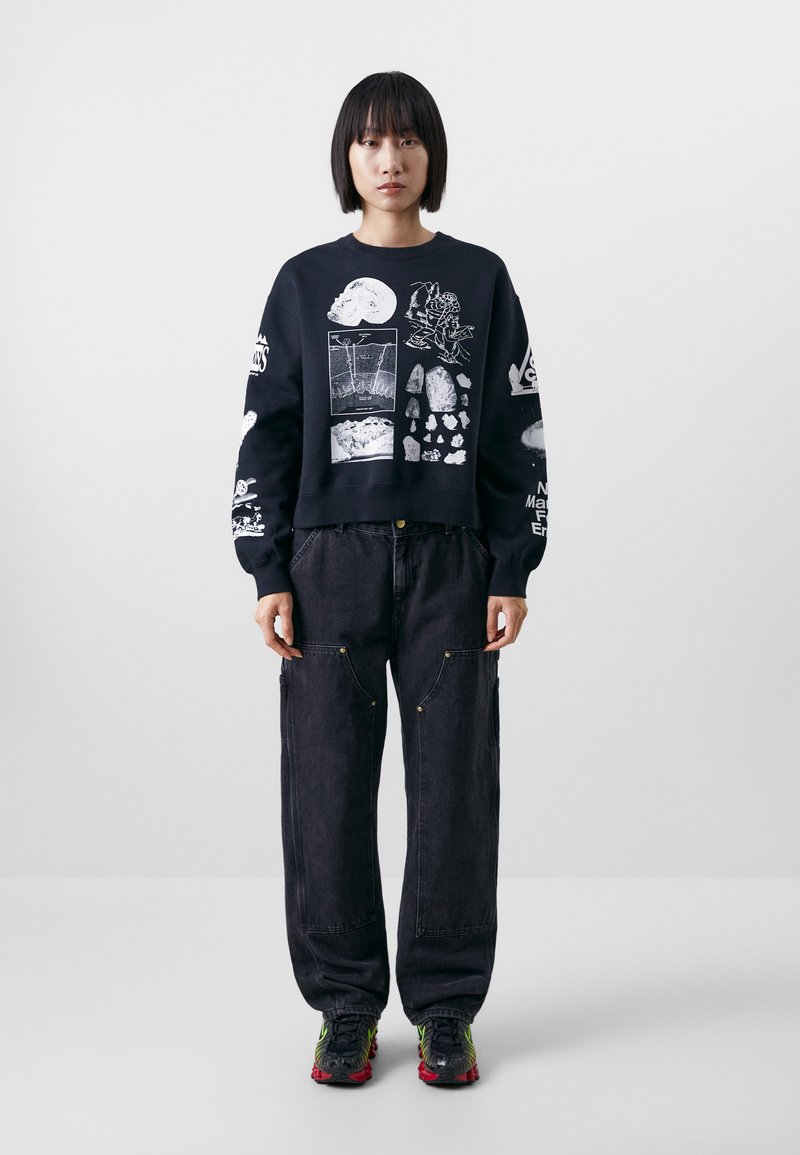 Felpa nera con stampe grafiche bianche, abbinata a pantaloni in denim scuro larghi con tasche laterali, completata da sneakers colorate.