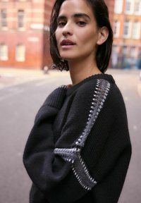 Schwarzer Strickpullover mit strukturierten grauen Akzenten an den Schultern, breitem Ausschnitt und lockerer Passform. Das Material wirkt weich und gemütlich.