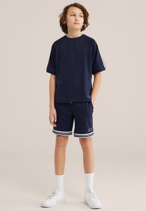 Chlapec s kučeravými vlasmi stojí a pozerá nahor, má na sebe tmavomodrý športový outfit s pruhovanými šortkami, biele ponožky a biele tenisky.