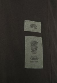 Deux étiquettes rectangulaires en tissu gris cousues sur un tissu foncé, présentant un petit texte noir avec les détails de la marque et du produit.