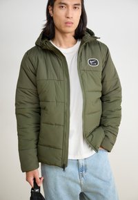 Žalias pūkuotas džemperis su kapuce, siuvinėtas dizainas, priekinis užtrauktukas ir baltas Nike AIR logotipas kairėje krūtinės pusėje. Dėvimas ant baltos marškinėlių.