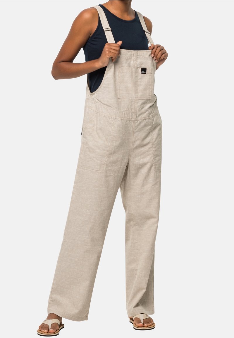 Beige linen overalls with wide legs, adjustable straps, and a front pocket; paired with a navy tank top and sandals.