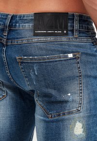 Jeans de mezclilla azul con un aspecto desgastado, que cuentan con una etiqueta de cuero negra, desvanecimiento claro y un parche cerca del bolsillo trasero como detalle.