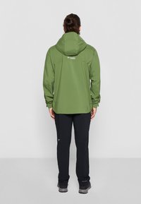 Chaqueta impermeable verde con capucha, que presenta un logo en la parte trasera. Combinada con pantalones negros y zapatos de senderismo grises. Textura suave y diseño ajustado.