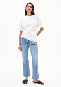 Weißes Baumwoll-T-Shirt mit kurzen Ärmeln, kombiniert mit hellblauen geraden Jeans. Das Outfit wird durch schwarze Slide-Sandalen ergänzt.